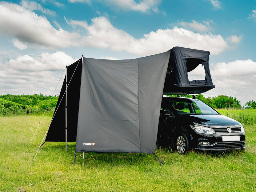 Small black car with the TentBox GO and Tunnel Awning set up in a grassy field under a blue sky with clouds.